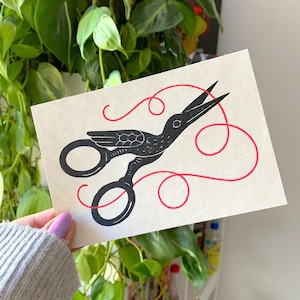 May include: A black and white art print featuring scissors with a bird design, cutting a red thread. The artwork is on a cream-colored paper, with a green plant in the background. The scissors have a unique, artistic design.