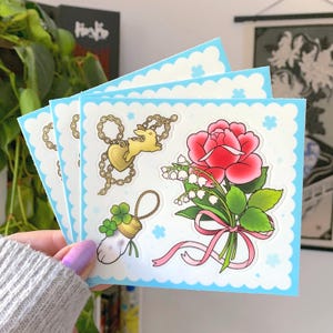 May include: A stack of stickers with a light blue scalloped border. The stickers feature illustrations of a red rose, a gold heart and bunny charm, a four-leaf clover, and a bouquet of lily of the valley with a pink ribbon.