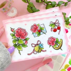 May include: A set of six pink and green floral stickers with a pink background. The stickers feature roses, a heart with a bow, forget-me-nots, cherries, and a letter.