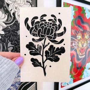 May include: Black and white linocut print of a single chrysanthemum flower with leaves. The flower is in the center of the print and is facing the viewer. The leaves are on either side of the flower.