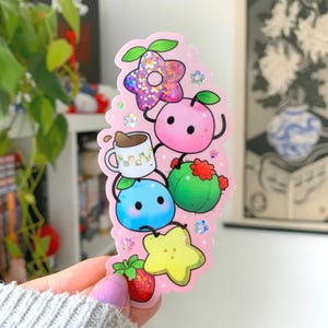 May include: A pink sticker featuring various cartoon food and plant illustrations. Includes a glittery star, a pink peach, a cactus, a blue blob, a strawberry, a yellow star, and a coffee mug. The sticker has a pink background with small white dots.