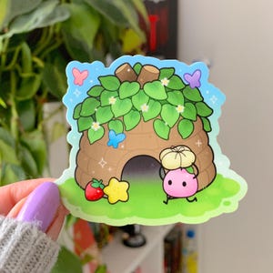 May include: A colorful sticker featuring a cartoon beehive with green leaves, a pink character, and a strawberry. The sticker has a blue sky background with butterflies and hearts.