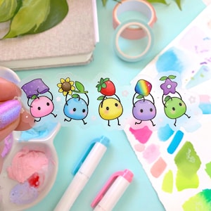 May include: A sheet of colorful stickers featuring cute cartoon characters holding various objects, including a sunflower, a strawberry, a rainbow, and a flower. The stickers are on a light blue background.