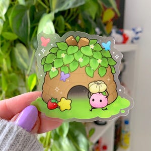 May include: A clear sticker featuring a cartoon illustration of a pink character carrying a yellow hat, walking towards a brown house with green leaves and butterflies. A strawberry and star are also included.