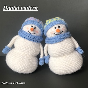 May include: Two crocheted snowmen with blue scarves and hats. One snowman has a pink and white hat, the other has a yellow and white hat.  The snowmen are standing on a gray surface.  Digital pattern.