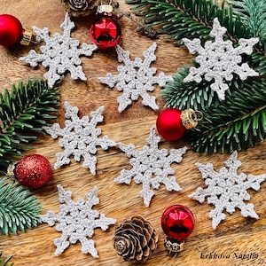 May include: A collection of crocheted snowflake ornaments in a light gray color, displayed on a wooden surface with red Christmas ornaments and pine branches.