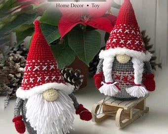 Peut inclure: Deux gnomes en crochet faits main avec des bonnets pointus rouges à motifs de croix blancs, des corps gris et des moufles rouges. Un gnome a une longue barbe en laine blanche, l'autre des tresses blanches. Le texte indique Christmas Gnomes Home Decor Toy et Crochet Pattern.