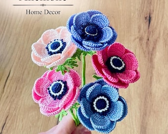 May include: A bouquet of five crocheted anemone flowers in shades of light pink, dark pink, and blue, each featuring a dark blue center with a white ring. The text Anemone Home Decor and Crochet Pattern is visible on the image.