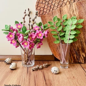 Pode incluir: Um buquê de flores de crochê rosa em um vaso de vidro transparente, ao lado de um buquê de folhas de crochê verdes em um vaso de vidro transparente. Há três ovos de codorniz em uma superfície de madeira.