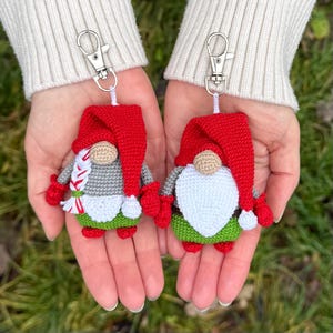 Pode incluir: Dois chaveiros de gnomos feitos à mão em crochê. Cada gnomo tem um chapéu vermelho, camisa cinza, barba branca e detalhes verdes e vermelhos. Os chaveiros têm fechos prateados e cordões brancos.