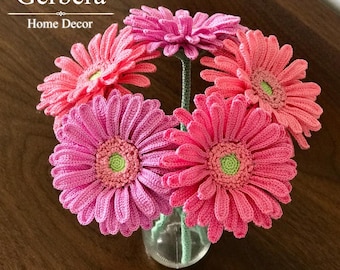 May include: A bouquet of five crocheted gerbera daisies in shades of pink and coral with light green centres, each on a thin green stem, arranged in a clear glass vase. The text Gerbera Home Decor and Crochet Pattern is visible.