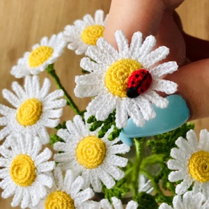 May include: Crocheted daisy ring featuring white petals, a yellow centre, and a small red and black ladybird. The ring is part of a collection of crocheted flowers, with green stems and leaves.