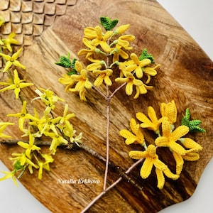 Könnte beinhalten: Zwei gehäkelte Zweige mit gelben Forsythienblüten und grünen Blättern auf einer Holzoberfläche. Der Text "Natalia Erkhova" ist in der linken unteren Ecke des Bildes sichtbar.