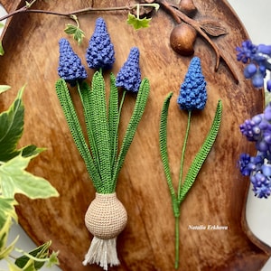 Pode incluir: Flores de jacinto-uva azuis de crochê com caules e folhas verdes. As flores são dispostas em um buquê e um único caule. O buquê tem uma base de bulbo de crochê bege com franjas brancas. Em uma bandeja de madeira.