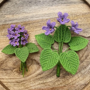 Op de afbeelding: Twee gehaakte viooltjesbloemstukken. Eén heeft een cluster van paarse bloemen, de andere heeft drie afzonderlijke bloemen. Beide hebben groene bladeren en stelen, tegen een houten achtergrond.