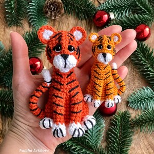 May include: Two crocheted tiger plushies, one larger than the other, with orange and black stripes. The larger tiger is sitting on a hand, while the smaller tiger is sitting in front of it. Both tigers have white faces and black eyes.