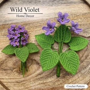 May include: Two crochet violet flower arrangements with purple petals and green leaves. The text Wild Violet Home Decor appears at the top, and the text Crochet Pattern appears at the bottom right. The flowers feature small yellow centres.