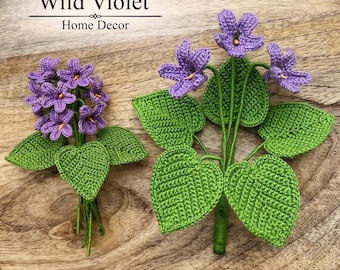 Puede incluir: Dos arreglos de flores violetas de ganchillo con pétalos morados y hojas verdes. El texto Wild Violet Home Decor aparece en la parte superior y el texto Crochet Pattern en la parte inferior derecha. Las flores tienen pequeños centros amarillos.