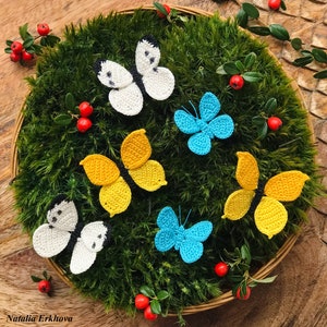 May include: A collection of crocheted butterflies in various colors, including yellow, blue, white, and black, displayed on a green moss background. The butterflies are arranged in a circular pattern, creating a whimsical and decorative display.