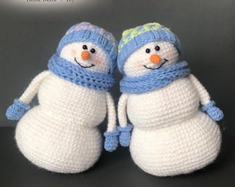 May include: Two white crocheted snowman figures with orange noses, black eyes, and pink cheeks. Each wears a blue scarf and blue mittens. One snowman has a blue and pink hat, while the other has a blue and yellow hat. Text reads Snowman Home Decor Toy.