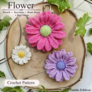 Op de afbeelding: Drie gehaakte bloemen: een grote roze bloem met een lichtgroen hart, een kleinere witte bloem met een geel hart en een middelgrote paarse bloem met een blauw hart. De roze bloem is voorzien van een sleutelhanger en een speld.