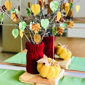 May include: A crocheted autumn-themed centrepiece with a red knitted vase, a yellow crocheted pumpkin, and a branch with crocheted leaves in shades of green, yellow, and brown.