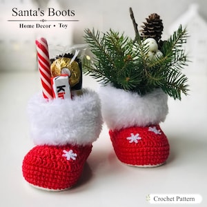 May include: Two red crocheted boots with white faux fur cuffs and small white snowflake appliques. One boot holds a candy cane and a chocolate bar, while the other contains pine branches, a pinecone, and white berries. Text reads Santa's Boots.