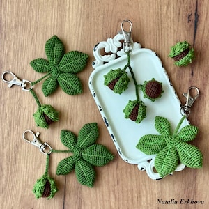 Può includere: Portachiavi all'uncinetto fatti a mano con foglie verdi e baccelli di castagno. I portachiavi hanno chiusure color argento. Le foglie e i baccelli sono disposti su un vassoio bianco e una superficie di legno.