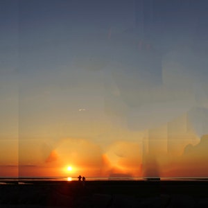 May include: Silhouettes of two people standing on a beach at sunset. The sky is a vibrant orange and yellow, with a few clouds.