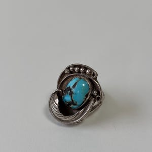 Könnte beinhalten: Ein silberner Ring mit einem großen, ovalen Türkisstein. Der Stein ist in einer verzierten Fassung mit Feder- und Perlendetails gefasst. Der Ring hat ein Vintage- oder antikes Aussehen.