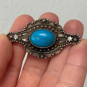Puede incluir: Un broche de plata con una piedra turquesa engastada en un diseño filigrano detallado.