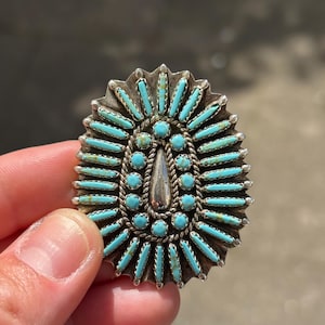 Könnte beinhalten: Ovaler silberfarbener Anhänger mit Türkissteinen. Der Anhänger hat eine zentrale Tropfenform und ist von einem Sonnenstrahl-Design aus Türkissteinen umgeben. Die Kanten haben ein gewelltes Design. Eine Hand hält den Anhänger.