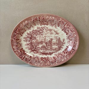 Könnte beinhalten: Ovale Servierplatte mit rot-weißem Design. Die Platte zeigt eine detaillierte Landschaftsszene mit einem Schloss und Bäumen in der Mitte, umgeben von einem floralen Rand. Ein klassisches Stück zum Servieren von Speisen.