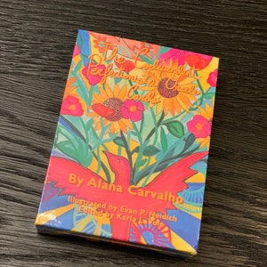 May include: A colorful box with the title "The Codependent Perfectionist's Oracle Cards" and the author's name "By Alana Carvalho". The box is illustrated with a red bird, sunflowers, and other flowers.