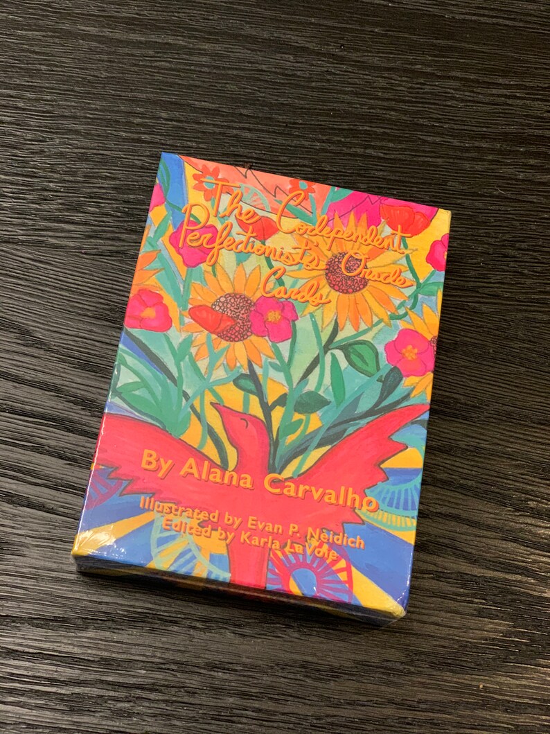 May include: A colorful box with the title "The Codependent Perfectionist's Oracle Cards" and the author's name "By Alana Carvalho". The box is illustrated with a red bird, sunflowers, and other flowers.