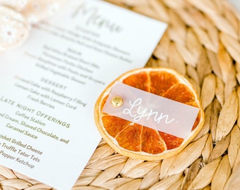 May include: A place setting with a woven placemat, a white card with a gold menu, and a dried orange slice with a white paper tag that says "Lynn".