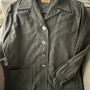 May include: A dark gray, long-sleeved jacket with a collared neckline and four buttons down the front. The jacket has two patch pockets on the front and a single pocket on the left sleeve.