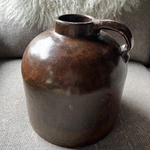 May include: A brown stoneware jug with a rounded body and a handle. The jug has a dark, glossy finish and a small opening at the top. The jug is likely an antique or vintage piece.
