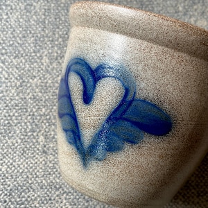 May include: A speckled ceramic mug with a blue heart design. The heart is stylized with wing-like accents. The mug has a neutral color palette with a brown rim.