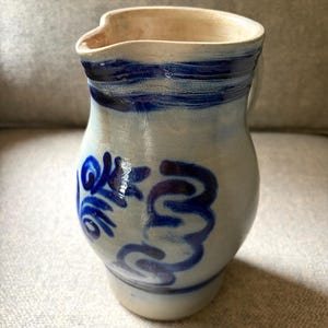 May include: A ceramic pitcher with a light gray, salt-glazed finish featuring hand-painted cobalt blue decorative swirls and floral patterns. The vessel has a wide, rounded body, a flared rim with a pouring spout, and a smooth, glossy surface texture.