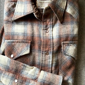 May include: A long-sleeve button-down shirt featuring a brown, tan, and grey plaid pattern. The shirt has a quilted brown interior lining, two chest pockets with flaps, and metallic snap buttons. A Pendleton brand label is sewn inside the collar.