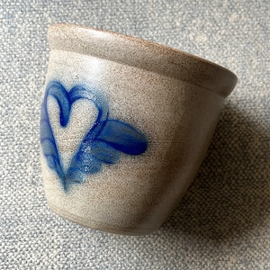 May include: A small, beige ceramic pot with a blue heart design. The pot has a speckled texture and a slightly angled rim. The heart is painted in a stylized, folk art style. This decorative pot is suitable for small plants or storage.
