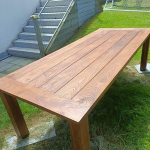 Puede incluir: Una mesa rectangular de madera con acabado natural, con tablero de lamas y patas cuadradas y robustas. La mesa está sobre un césped, lo que sugiere uso en exteriores. La veta de la madera es visible.