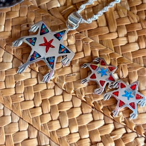 Puede incluir: Un collar con colgante de plata y pendientes a juego con un diseño de estrella con una estrella roja en el centro y detalles coloridos. Las joyas se exhiben sobre una superficie de paja tejida.