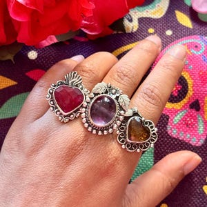 Peut inclure: Trois bagues en argent portées sur les doigts. La bague de gauche présente une pierre rouge en forme de cœur avec un sommet en forme de flamme. La bague centrale a une pierre ovale violette entourée de feuilles. La bague de droite est ambrée.