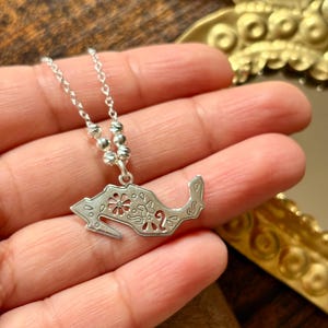 Puede incluir: Un collar plateado con un colgante detallado. El colgante presenta un diseño floral con detalles recortados. El collar se sostiene en una mano, mostrando el intrincado diseño y la artesanía. La cadena es delicada, lo que aumenta la elegancia del collar.