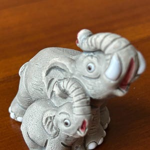 May include: A gray ceramic figurine of two elephants, one adult and one baby, nestled together. The elephants have white tusks and red accents on their mouths and ears. The figurine is detailed and sits on a wooden surface.
