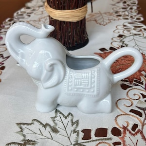 May include: A white ceramic elephant creamer with a trunk spout and a handle. The elephant has detailed features, including large ears and a textured saddle design. The creamer is a decorative kitchenware item.