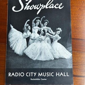 May include: A vintage black and white program cover for "Showplace" at Radio City Music Hall. The cover features a group of ballerinas in white dresses and tiaras. The text "Showplace" is in a cursive font.