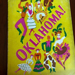 May include: A vintage yellow poster with the word "OKLAHOMA!" in pink and white. The poster features colorful illustrations of dancing figures in cowboy and western attire, with a brown wooden background.